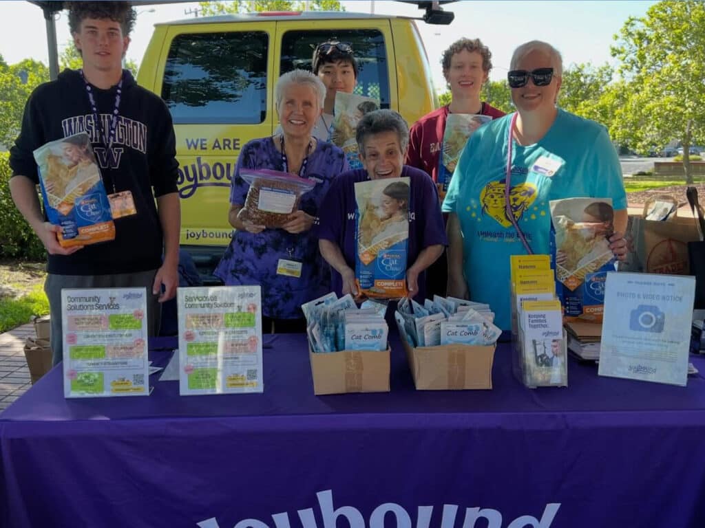 A group of five volunteers holding food packages stands behind a table covered with a purple cloth featuring the text "Joybound." Informational flyers are displayed on the table, with a van in the background and trees visible in the outdoor setting.
