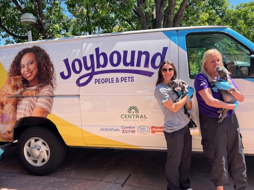 Two women stand outside a van marked "Joybound People & Pets," holding small dogs. One woman has curly hair and wears sunglasses, while the other has straight hair and wears a purple shirt. The van has colorful branding and logos from associated organizations.