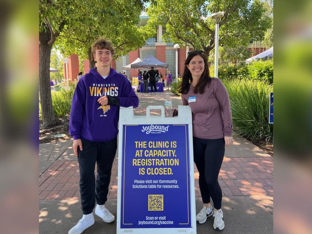 A young man and a woman stand beside a sign indicating the clinic is at capacity and registration is closed. The man wears a purple Minnesota Vikings sweatshirt, while the woman wears a light purple top. Trees and a building are visible in the background.