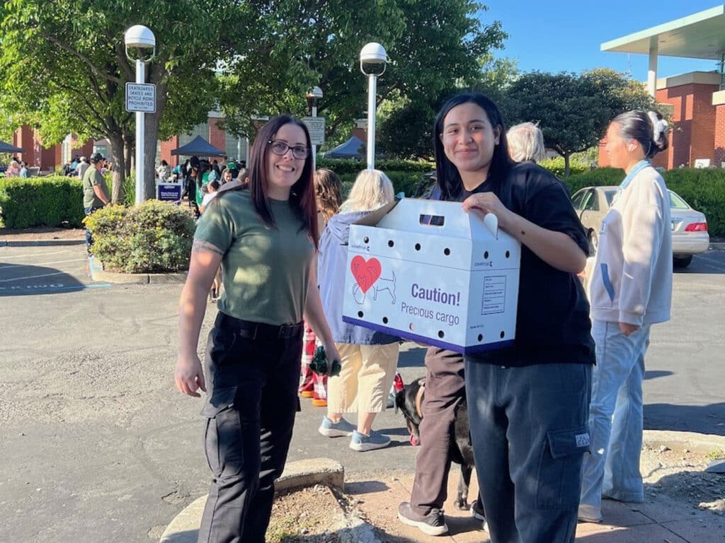 Two women stand outside a building, smiling while holding a box labeled "Caution! Processed Cargo". In the background, people can be seen participating in an event, with trees and vehicles visible.