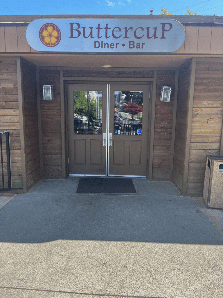 The entrance of ButtercuP Diner Bar features a double door with large glass windows, framed in light brown wood. Above the door is a sign with the name 'ButtercuP' in large letters, along with the words 'Diner Bar,' set against a circular logo.