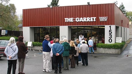 A group of people waiting in line in front of a store named 'The Garret Thrift Shop' with a brown and white exterior. The line extends along the building, and there are several cars parked nearby, indicating a busy event.