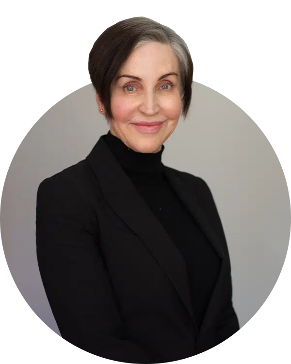 A woman with short dark hair and gray highlights smiles while wearing a black blazer over a black turtleneck. The background is a neutral gray, emphasizing her facial features.