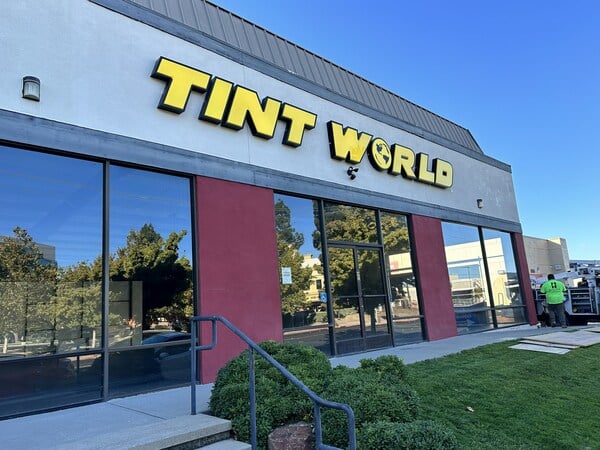 The exterior of a business building featuring a sign that reads "TINT WORLD" in large yellow letters. The façade includes windows reflecting the surrounding trees and grass, with a pathway leading to the entrance and a worker in a green shirt standing nearby.