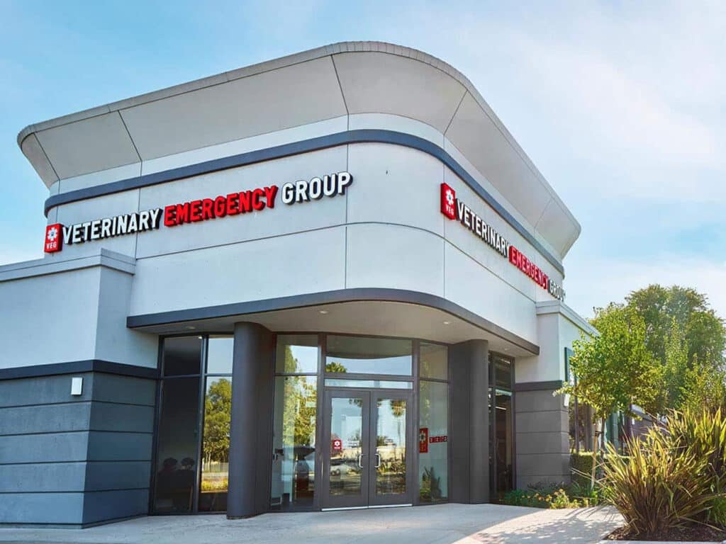 The exterior of the Veterinary Emergency Group building, featuring a modern architectural design. The sign displays the name 'Veterinary Emergency Group' prominently on the front of the building. Green landscaping and trees are visible around the entrance.