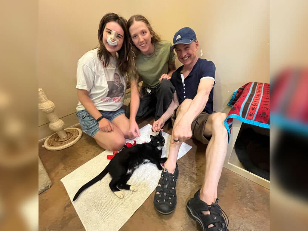 Three people sitting on the floor of a room with neutral-colored walls, interacting with a black cat. One person has a bandage on their nose, and they are all smiling. A scratching post and a colorful blanket are visible in the background.