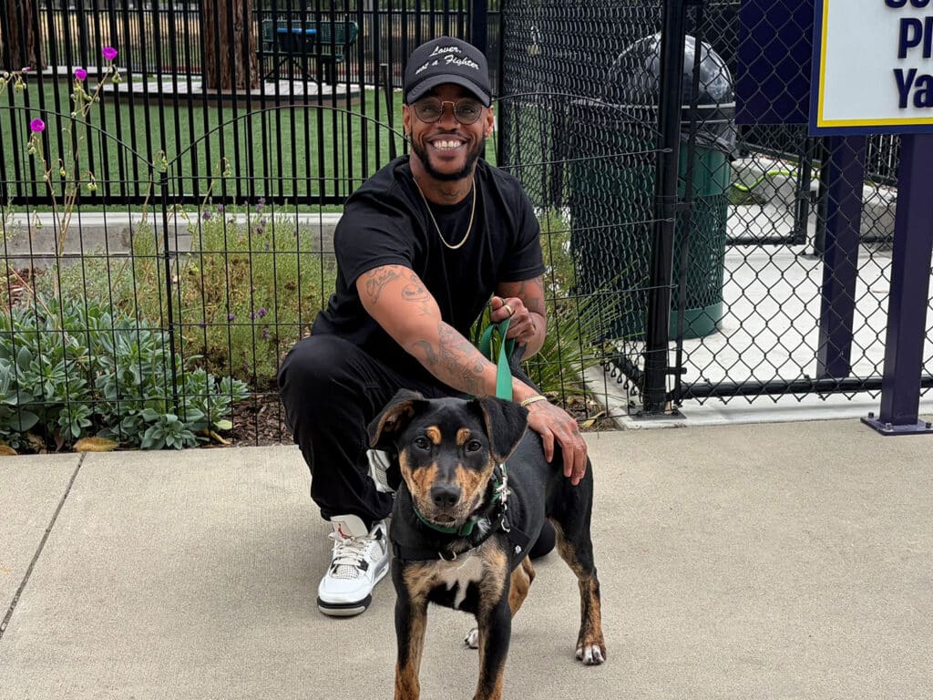 A man with glasses and a black cap squats next to a medium-sized dog with a black and tan coat. They are outside a fenced area with green grass and a recycling bin visible in the background.