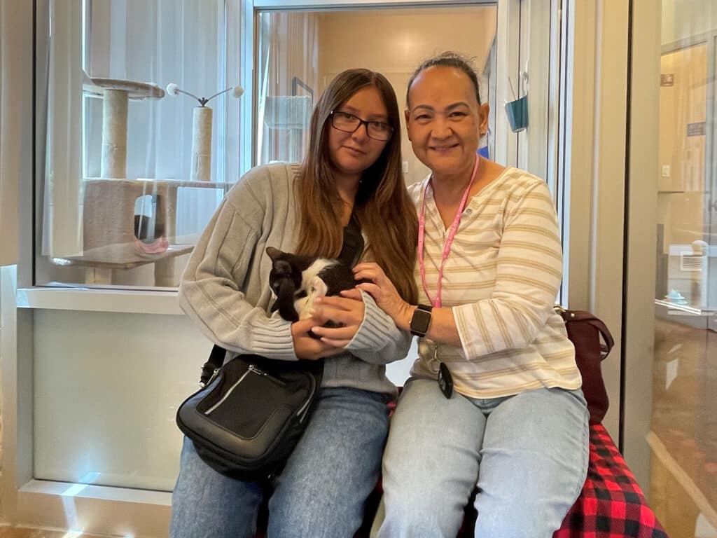 A young woman in a gray sweater holds a small black and white puppy while sitting next to an older woman in a striped shirt. They are both seated on a red and black checkered blanket in a well-lit room with cat towers in the background.