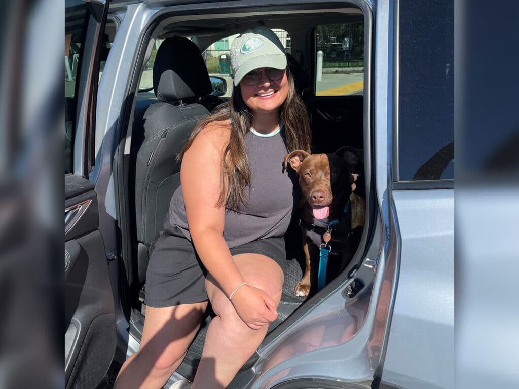 A woman with long dark hair, wearing a gray tank top and baseball cap, sits in the driver's seat of a car. Next to her is a light-brown dog wearing a black harness, both looking towards the camera. The image suggests a sunny day.