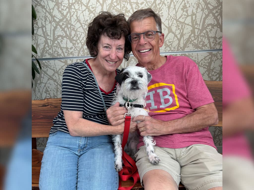 A couple sits on a wooden bench, each with a hand on a small dog in between them. The woman wears a striped black and white shirt and light jeans, while the man wears a pink shirt with a logo and light shorts. The background is softly patterned.