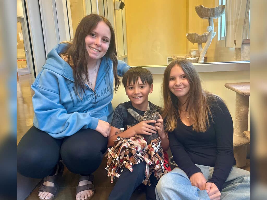 A young boy holds a kitten while sitting on the floor between two smiling young women. One woman wears a light blue hoodie, and the other has long brown hair. The background features natural light and cat trees in a room.