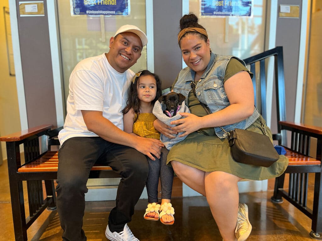 A man and woman sit on a bench with a young girl between them, all smiling. The woman holds a small black puppy. The background features windows and a sign that reads "Best Friend." The setting looks like an animal adoption center.