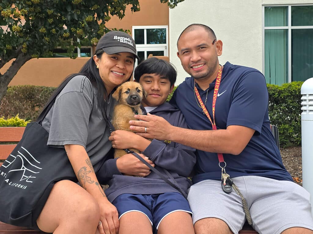 A woman in a grey shirt and a black tote, a boy in navy shorts, and a man in a black polo shirt pose on a bench. The boy holds a small brown puppy, while the man has his arm around him. In the background, there are green trees and a building.