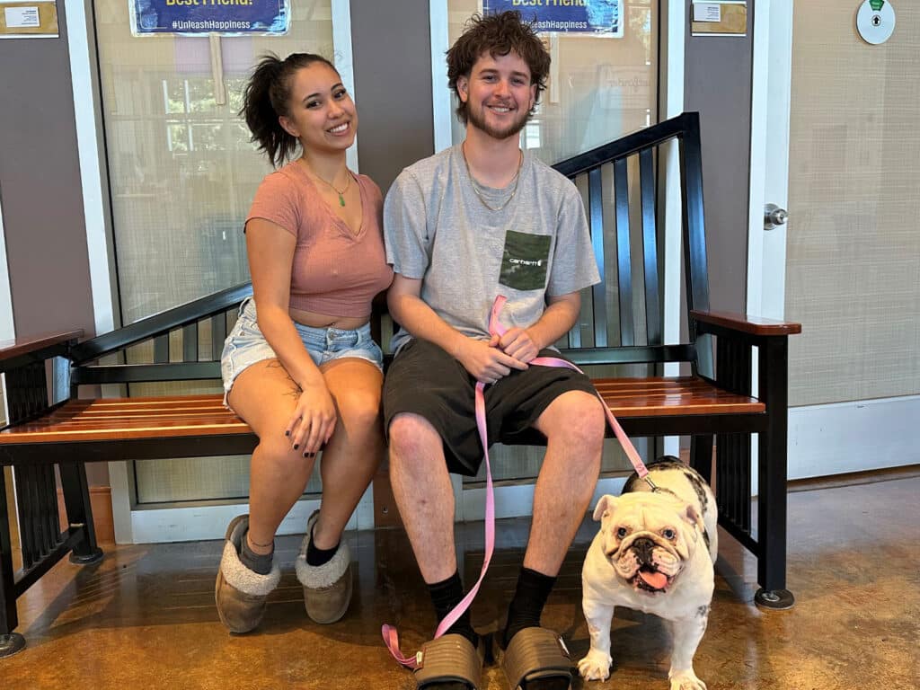 A young woman with long dark hair sits on a bench next to a young man wearing a gray t-shirt and black shorts. They are both smiling at the camera while a white bulldog with brown patches sits in front of them on the floor, leashed with a pink collar.