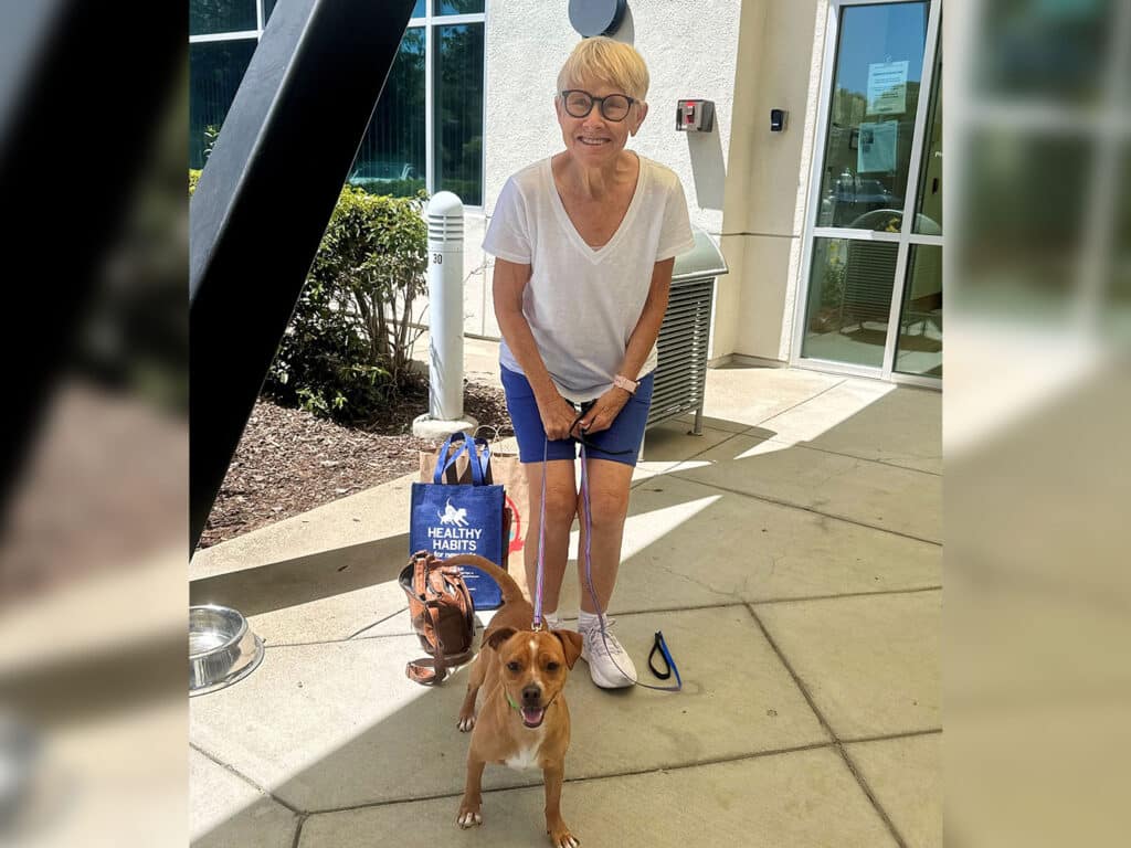 A woman wearing a white v-neck shirt and blue shorts holds the leash of a small brown dog while standing outside a building. A second brown dog is sitting beside her. She carries a blue bag with the text "Healthy Pets."