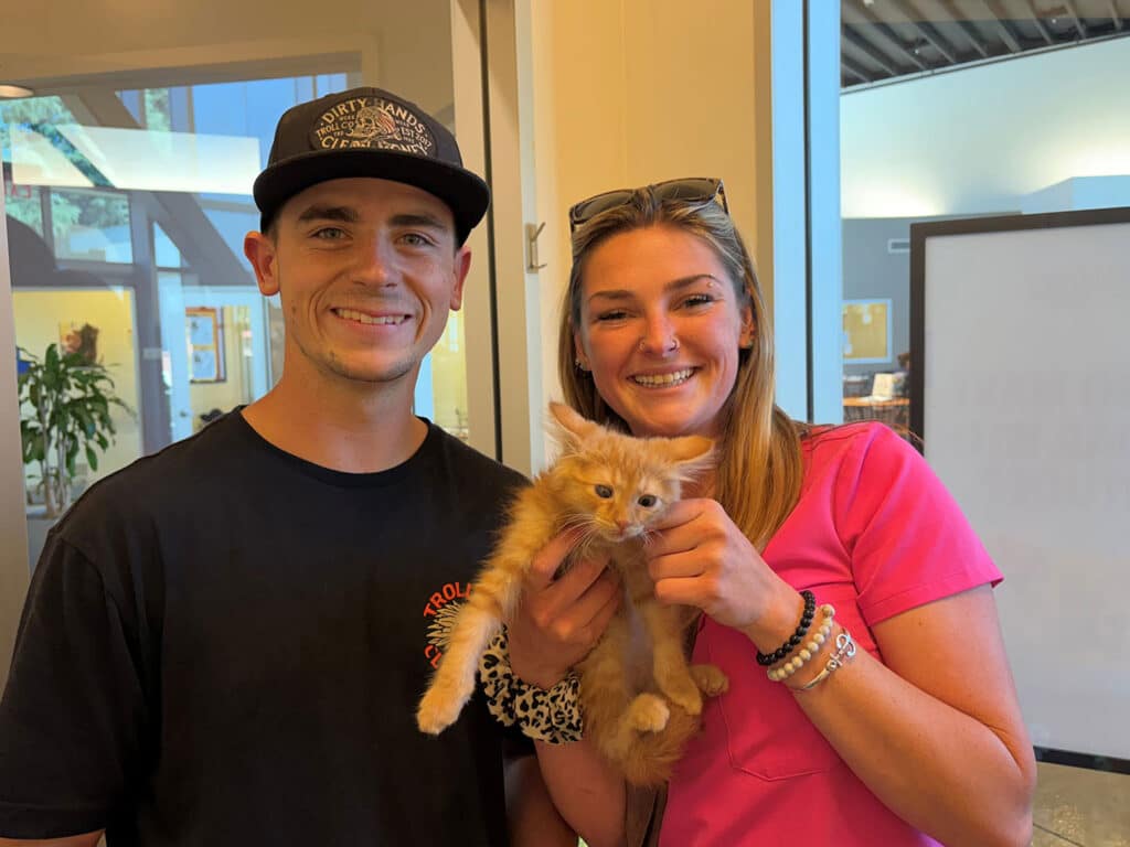 A young man in a black t-shirt and a cap stands next to a smiling woman wearing a pink shirt. The woman holds an orange kitten in her arms, while both individuals are positioned indoors with a light-colored backdrop.