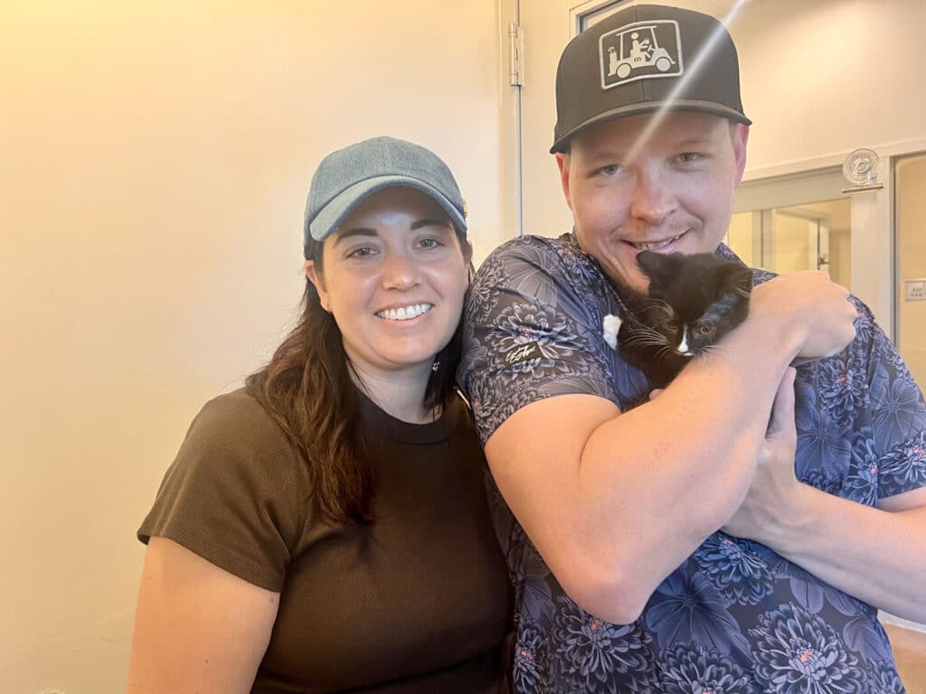 A woman wearing a dark shirt and a blue baseball cap stands next to a man in a blue patterned shirt and black hat. The man holds a black and white kitten in his arms. The background features light-colored walls and a door.