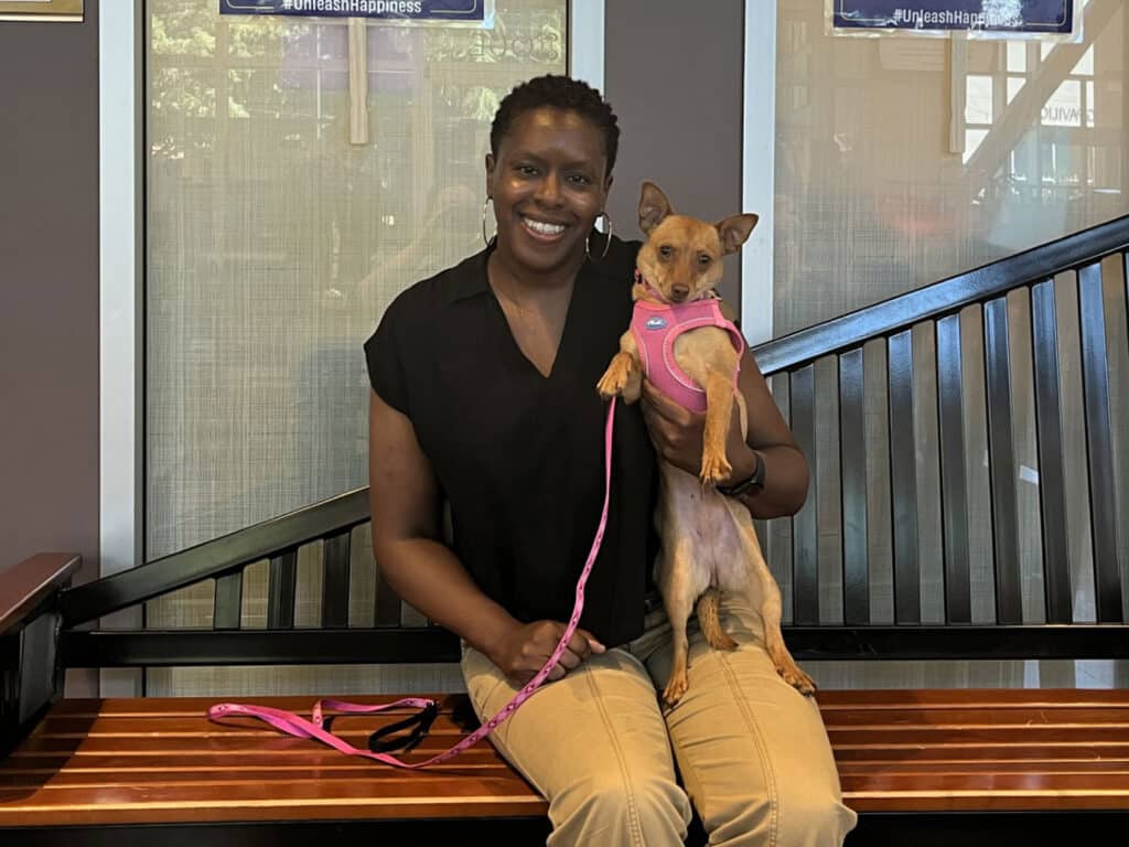A woman with braided hair wearing a black blouse smiles while sitting on a wooden bench, holding a small tan dog with a pink harness. The background shows a staircase and a soft-lit indoor setting.