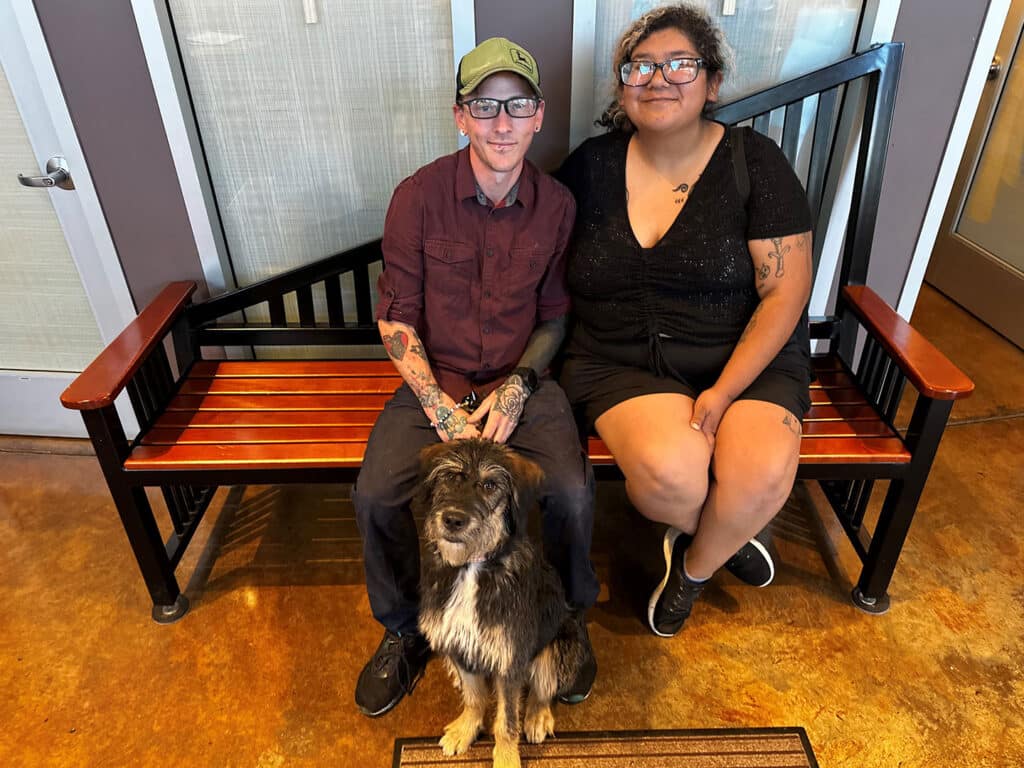 A man with glasses and tattoos sits on a wooden bench beside a woman with short curly hair. They are both wearing casual clothing, and a medium-sized dog with a shaggy coat sits in front of them on the bench.