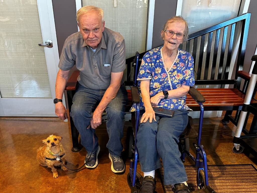 An older man wearing a gray shirt and dark jeans sits next to an elderly woman in a patterned blue shirt, seated in a wheelchair. A small brown dog on a leash sits beside the man. They are indoors, with a modern backdrop.