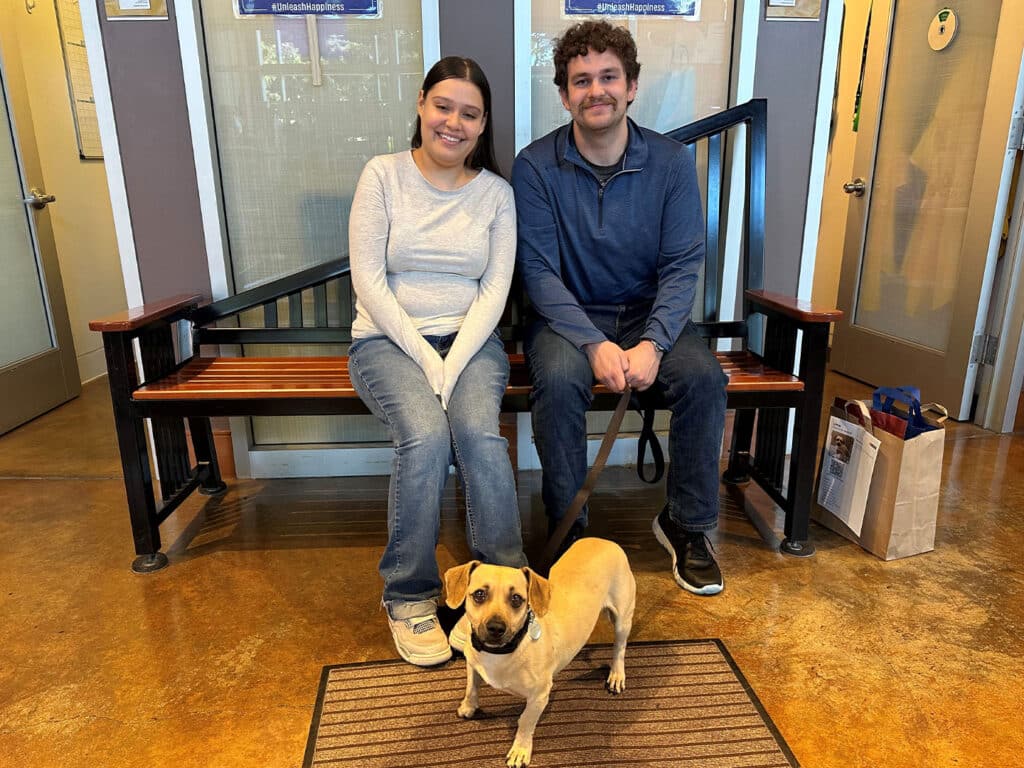 A young woman in a gray sweater and a young man in a blue sweater sit on a wooden bench inside a building. A small tan dog with a collar is sitting in front of them on a mat, with a bag visible nearby.