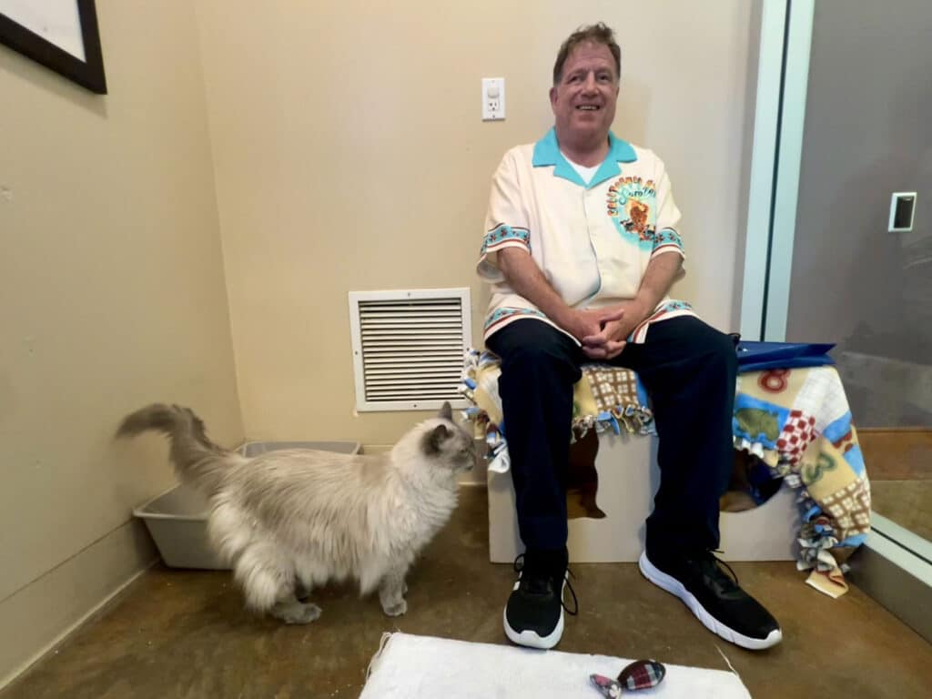 A man in a colorful shirt sits on a white box, smiling. Beside him, a light gray cat approaches on a concrete floor. The background shows a glass wall and a blanket with a colorful pattern draped over the box.
