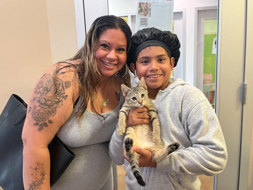 A woman with long hair, wearing a gray dress and holding a black bag, stands next to a boy in a gray sweatshirt and a black cap. The boy holds a gray tabby kitten in his arms, smiling at the camera. The background shows a doorway and colorful decorations.