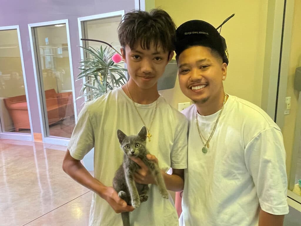 A young man in a white t-shirt holds a gray kitten, while another man in a white t-shirt and black cap stands beside him. They are inside a brightly lit space with large windows and plants in the background.