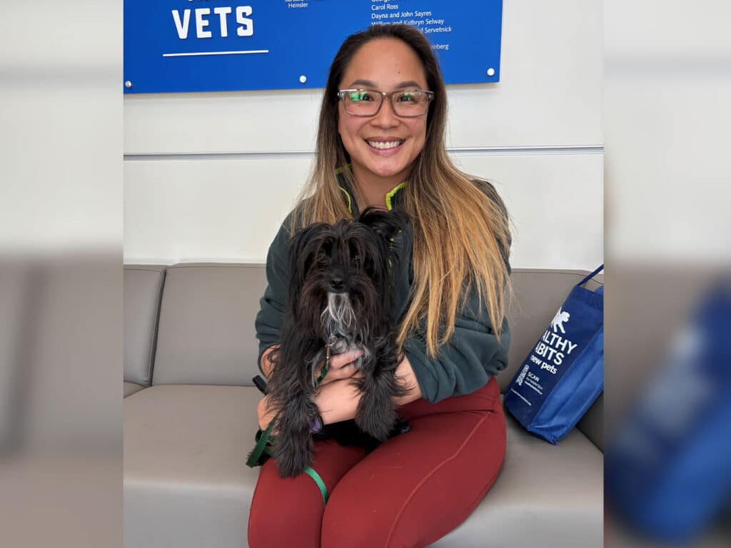A woman with long brown hair, wearing glasses and a dark green sweatshirt, sits on a gray couch holding a small black dog. The background features a blue sign with the text 'VETS'.