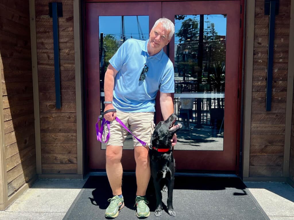 A man wearing a light blue shirt and beige shorts stands outside a building with large wooden doors. He holds a purple leash attached to a black dog with a red collar, both looking toward the entrance.