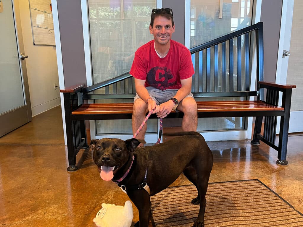A man wearing a red t-shirt is seated on a wooden bench with a black dog standing beside him. The dog is on a leash, looking toward the camera. The background shows a modern interior space with large windows.