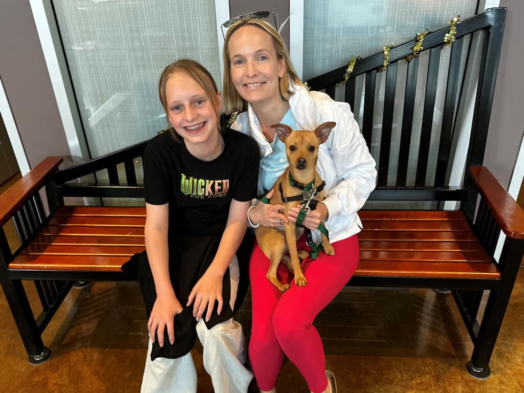 A woman wearing a white jacket and red pants sits on a wooden bench, holding a small dog on her lap. Beside her, a young girl in a black t-shirt with the word 'Wicked' sits with her arm around the woman, both posing for the photo.