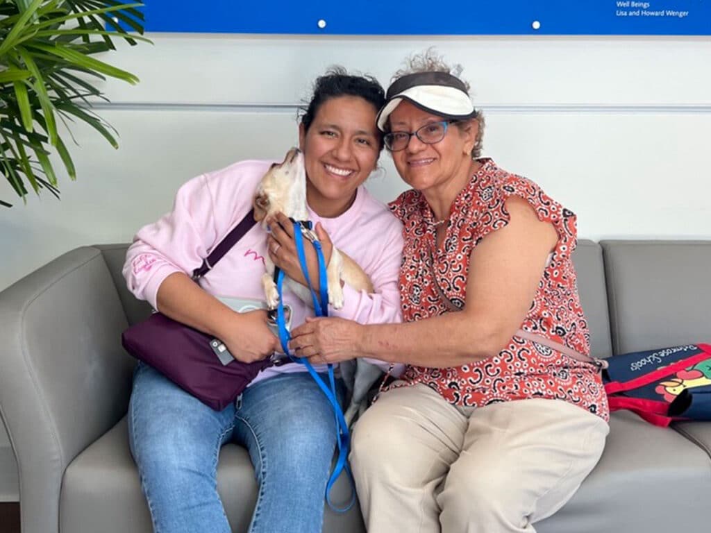 A young woman wearing a pink sweatshirt holds a small dog while sitting next to an older woman in a red patterned blouse. Both are seated on a gray couch in an indoor setting with plants and wall decor visible in the background.