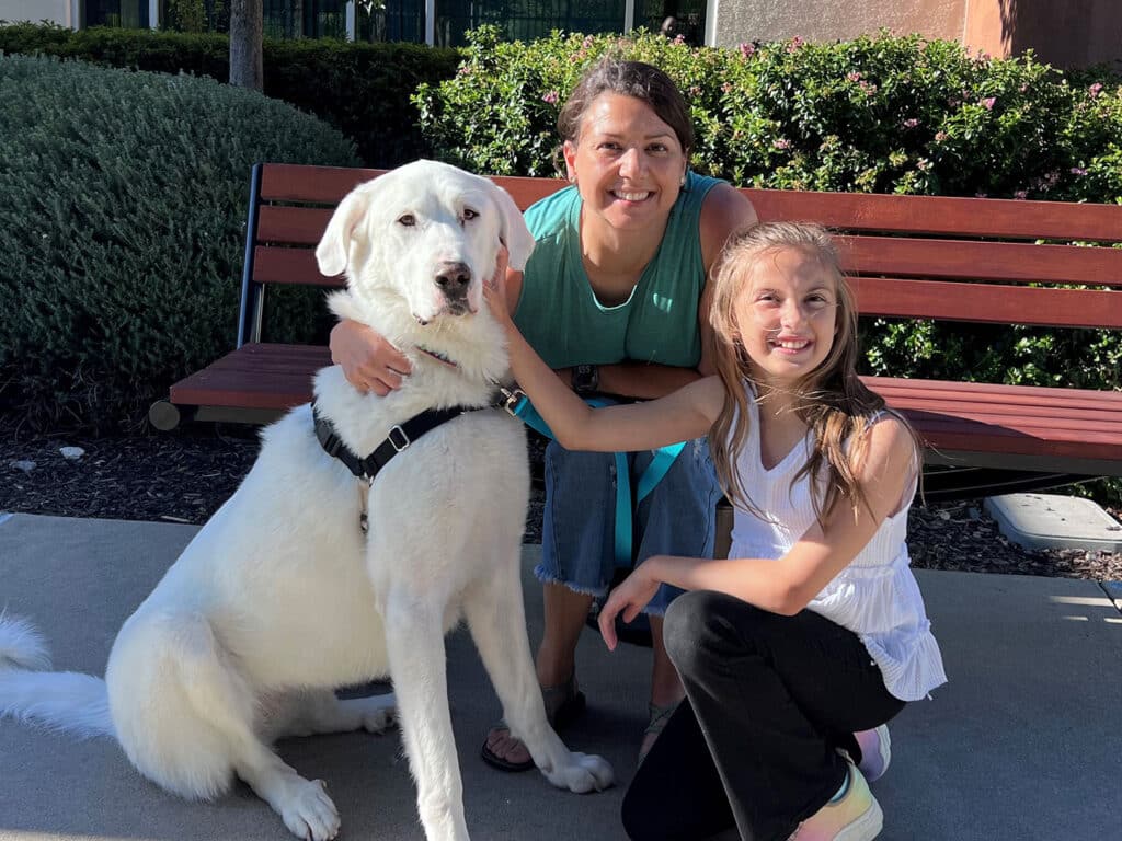 A woman with dark hair in a teal tank top smiles while sitting next to a large white dog with a black harness. A girl in a white tank top sits beside them, both posing outdoors near a bench and greenery.