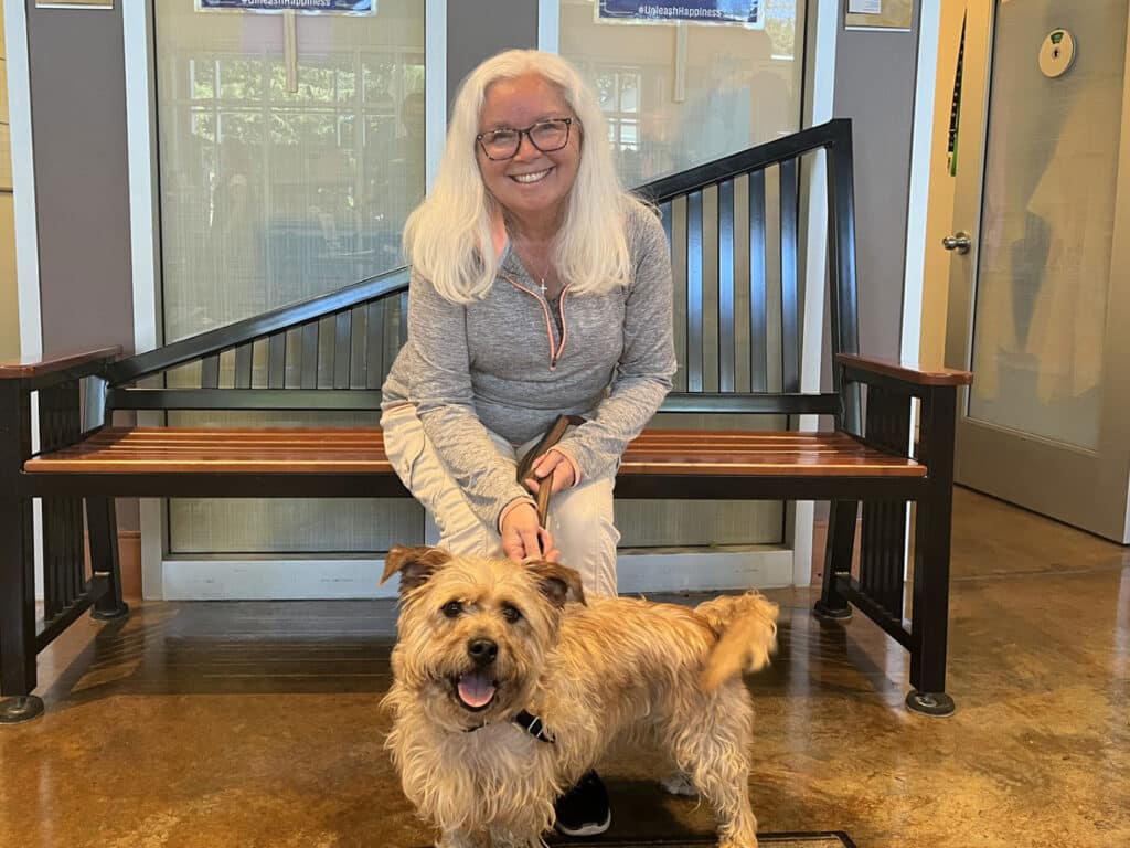 A woman with long, white hair and glasses sits on a bench, holding the leash of a small, tan dog. The setting appears to be a spacious indoor area with large windows and natural light.