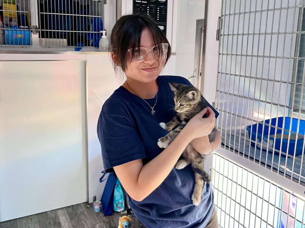A young woman wearing a dark t-shirt and eyewear holds a gray tabby kitten in her arms. The background includes a large pet enclosure with multiple compartments and items for animal care.