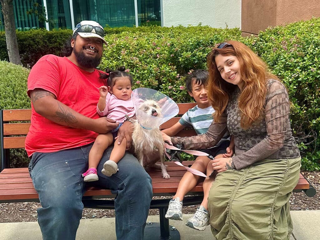 A family of four sits on a wooden bench in a garden area. The father, wearing a red shirt and sunglasses, holds a young girl in a pink dress. The mother, in a gray top and skirt, sits beside their son, who plays with a small dog wearing a cone collar.