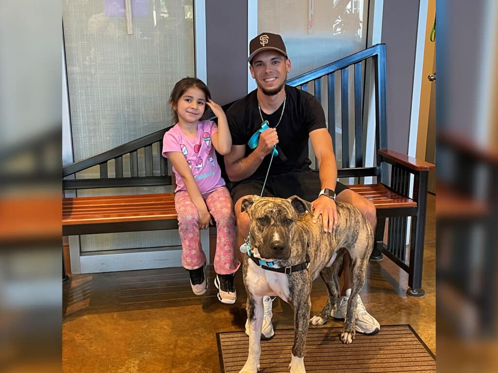 A young girl sitting on a bench next to a man, both smiling. The girl wears a pink shirt and pajama pants. They are accompanied by a gray dog on a leash. The background features a blue railing and a well-lit indoor space.