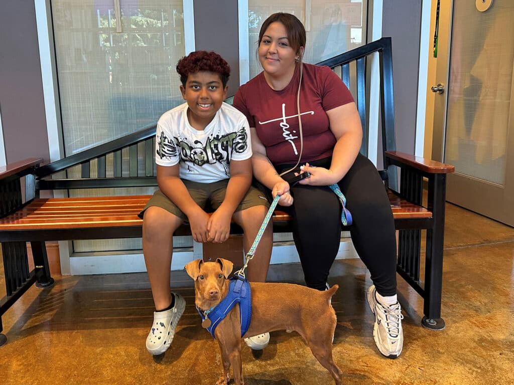 A young boy with curly hair wearing a white shirt and shorts sits on a bench next to a woman in a red shirt. A small brown dog on a leash sits in front of them. The setting features a modern indoor space with large windows.
