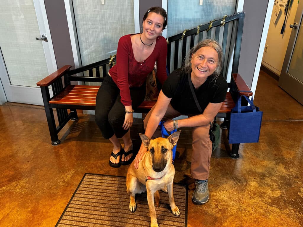 A young woman in a red top and black pants sits on the edge of a wooden bench next to an older woman wearing a black shirt and tan pants. A dog sits in front of them on a welcome mat inside an adoption center with large windows in the background.
