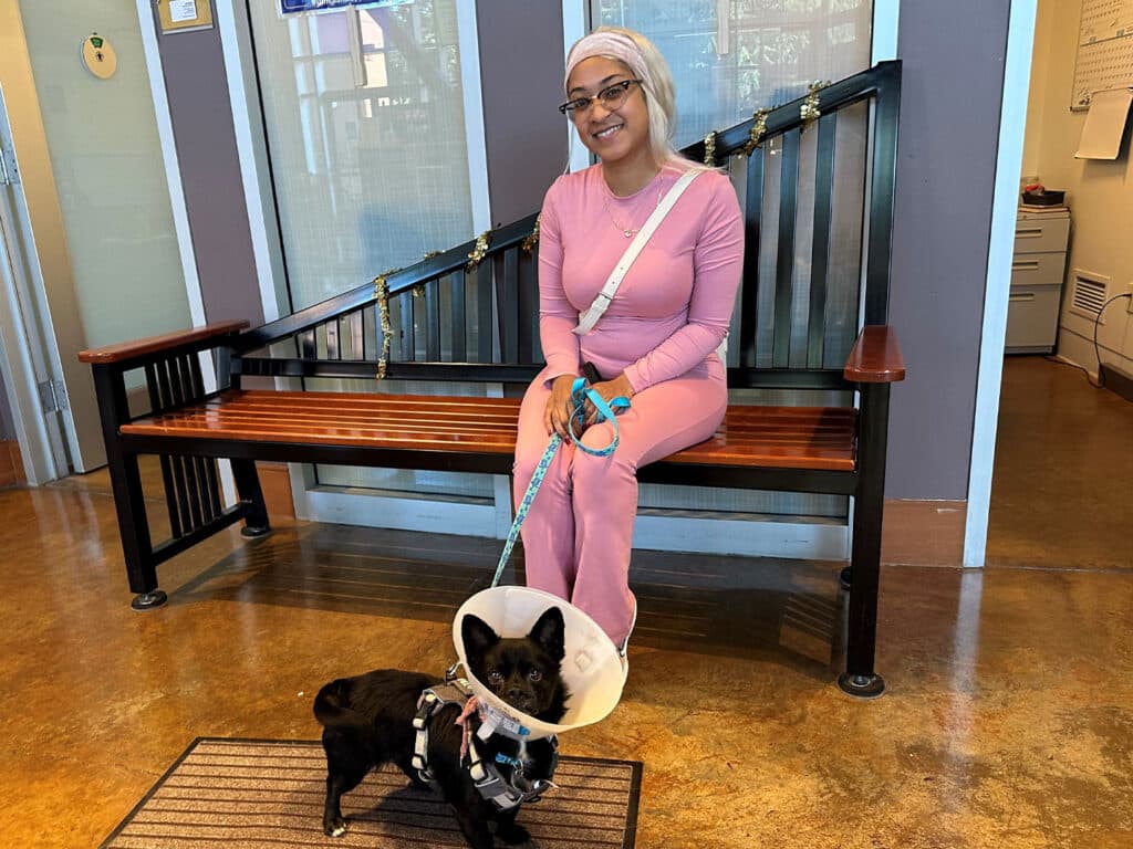 A woman in a pink outfit sits on a bench inside a building, holding the leash of a small black dog wearing an Elizabethan collar. The dog stands on a mat in front of her, with a welcoming interior visible behind them.