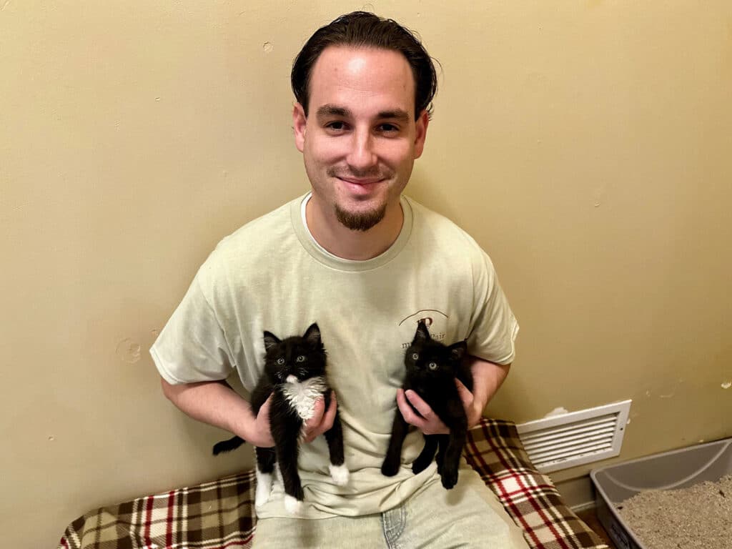 A young man with short brown hair wearing a light green shirt holds two black kittens, one in each arm. The man is seated on a plaid blanket, and the background shows a wall with an air vent and a litter box.