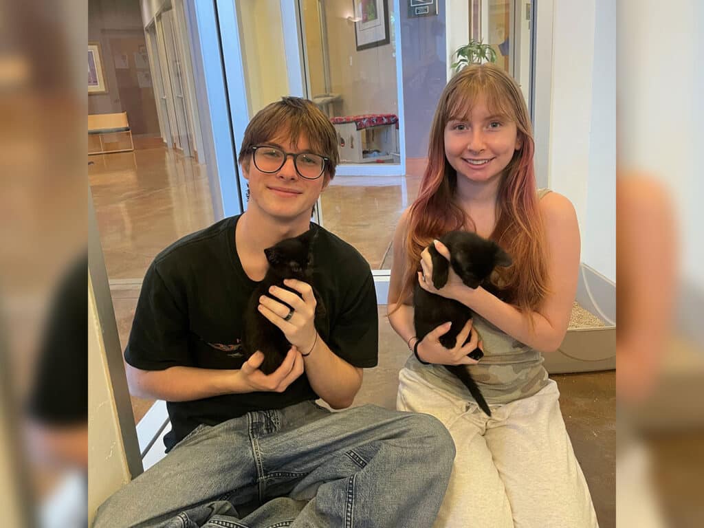 A teenage boy wearing glasses and a black shirt sits on the floor, holding a small black kitten. Next to him, a teenage girl with long hair, wearing a light tank top, also holds a black kitten. They are in a well-lit indoor space.