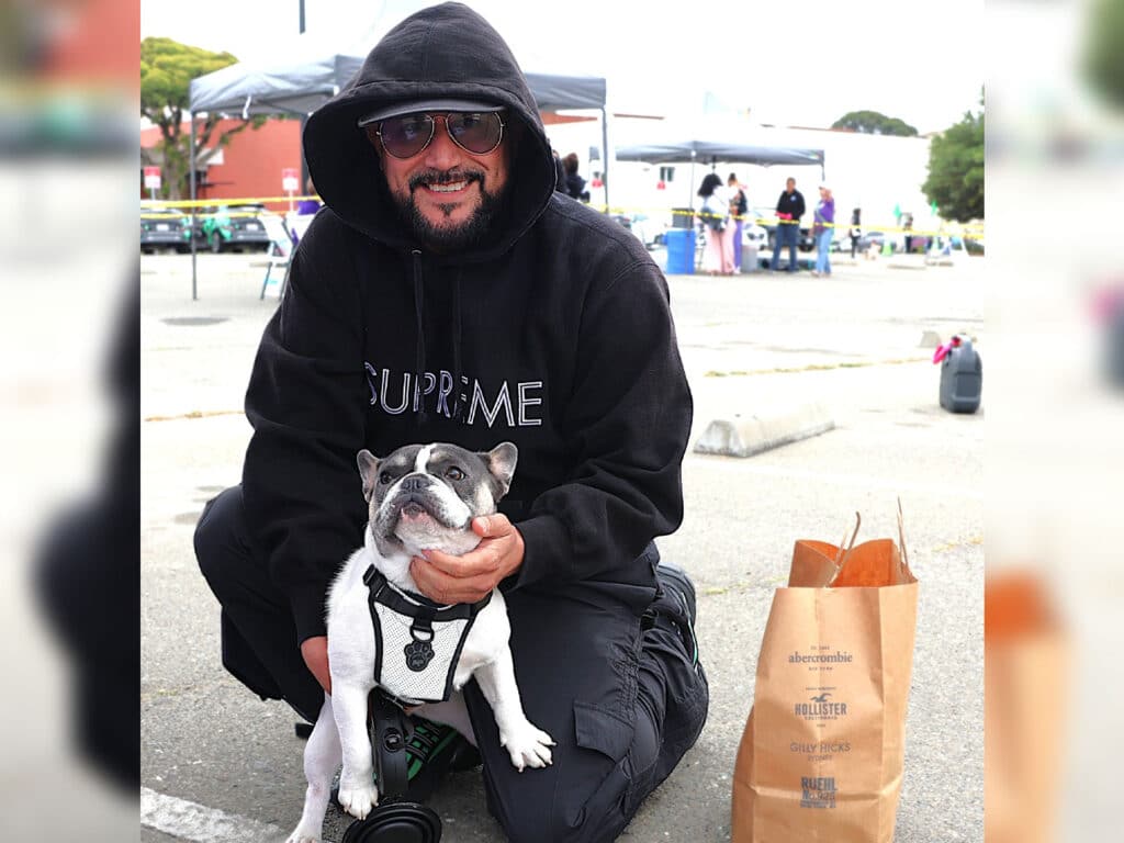 A man wearing sunglasses and a black hoodie with the word "SUPREME" smiles while kneeling next to a small white dog with black markings. A paper bag is visible on the ground beside them, with a blurred scene of a community event in the background.