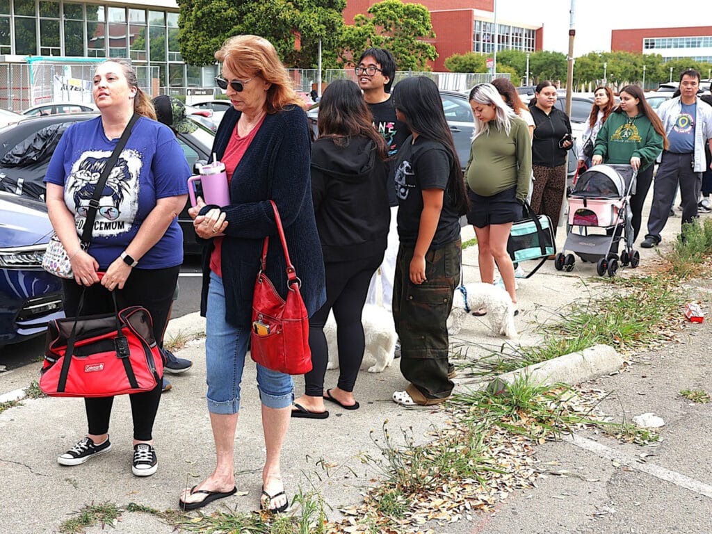 A group of people wait in line outside a building, some carrying bags and strollers. The crowd includes individuals of varying ages, with a few dogs present, while trees and a red-brick building are visible in the background.
