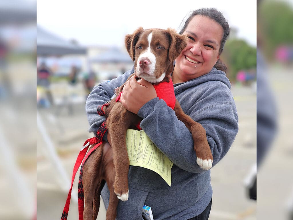 A woman in a gray hoodie holds a brown and white puppy in her arms. She is smiling at the camera, and a document is visible in her other hand. The background includes tents and outdoor space.