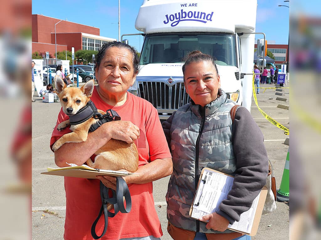 A woman in a red shirt holds a small dog while standing next to another woman wearing a gray vest. Both are in front of a Joybound truck, with people and tents visible in the background.