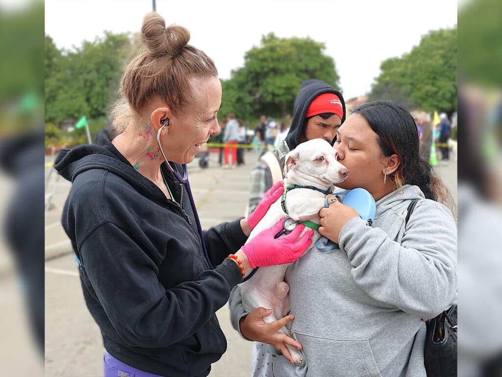 A woman wearing a black jacket and pink gloves holds a small white dog, while another woman in a gray hoodie leans in to kiss the dog. A crowd and green trees are visible in the background.