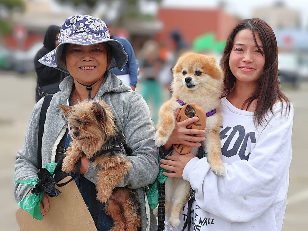 A woman wearing a patterned hat and gray jacket holds a small dog with a black harness. Beside her, another woman in a white sweatshirt holds a Pomeranian dog, both smiling in an outdoor setting with blurred figures in the background.