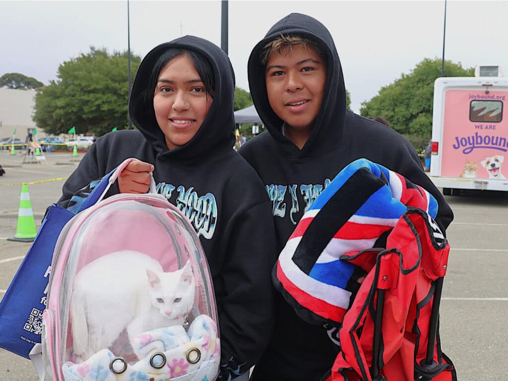 A young woman wearing a black hoodie and a young man wearing a black hoodie stand together. The woman holds a clear pet carrier with a white cat inside, while the man carries a red-and-black backpack. A food assistance vehicle is visible in the background.